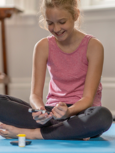 A young girl and her dog sit on a yoga mat as she uses a glucose monitor. 